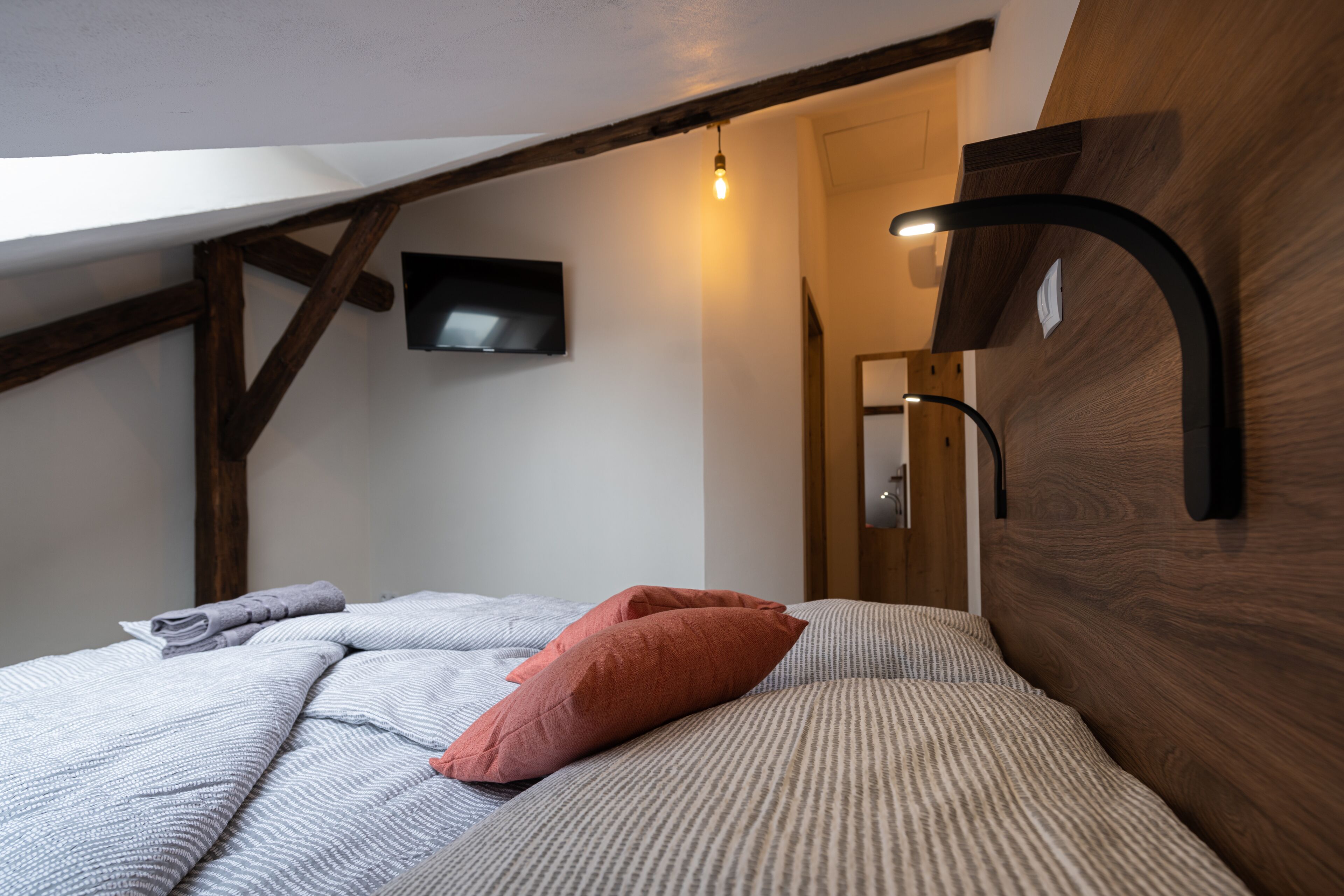 A bedroom with a sloped ceiling, a bed with striped bedding, a red pillow, a mounted TV, and a wall-mounted light fixture.