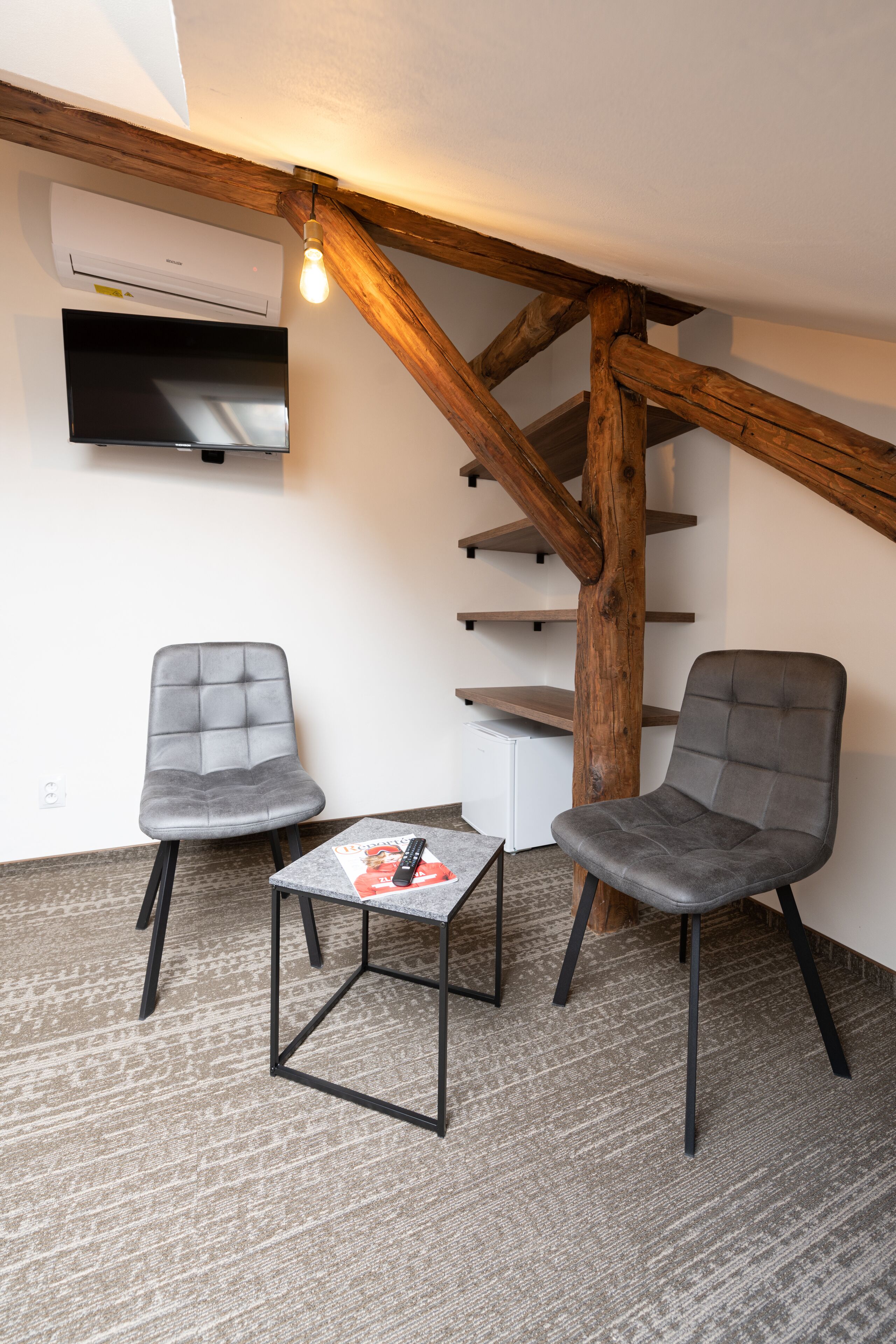 A room with two grey chairs, a small table with a magazine, a TV mounted on the wall, and a wooden staircase.