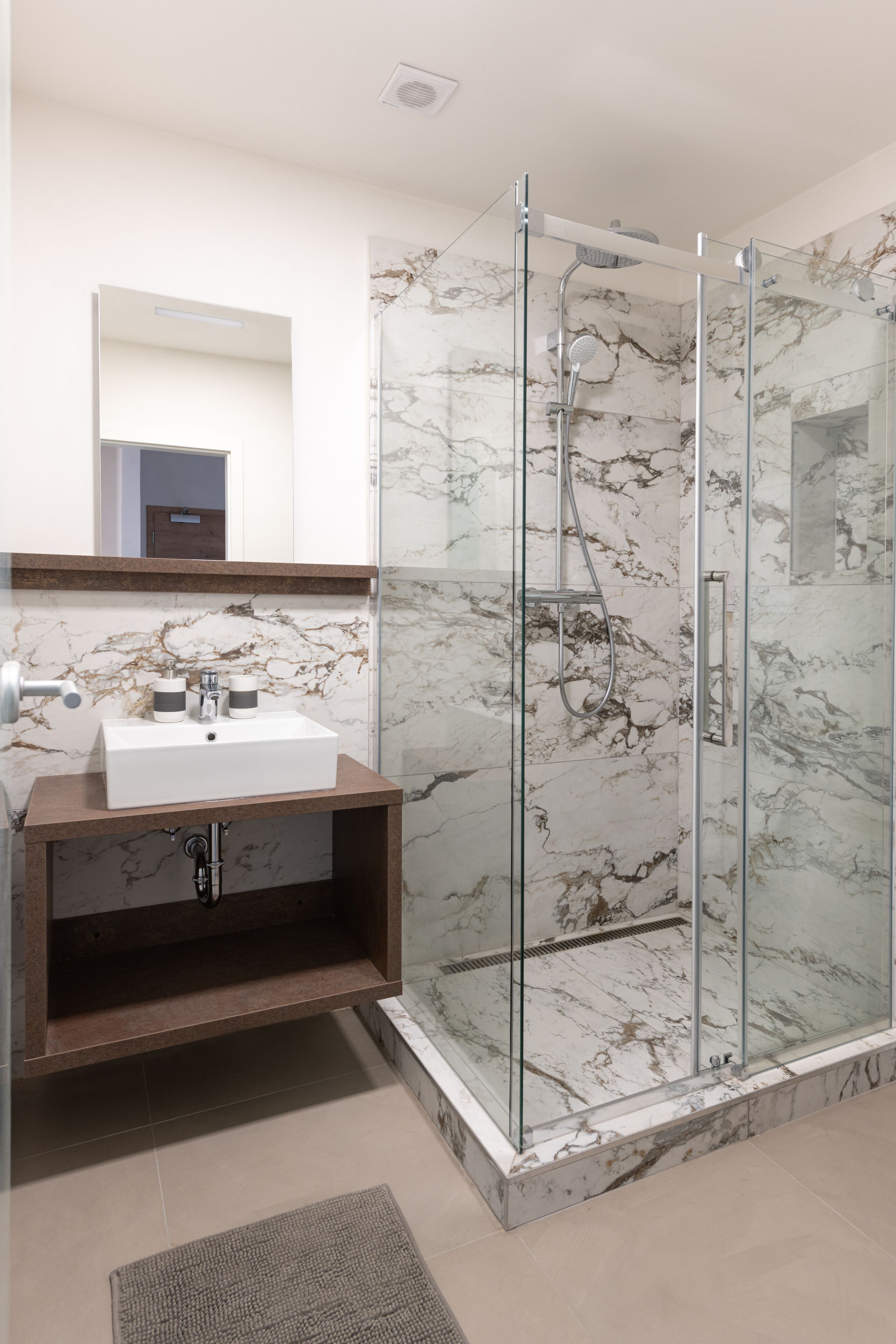 Un baño moderno con ducha de vidrio, pared con azulejos de mármol y tocador de madera con lavamanos blanco.