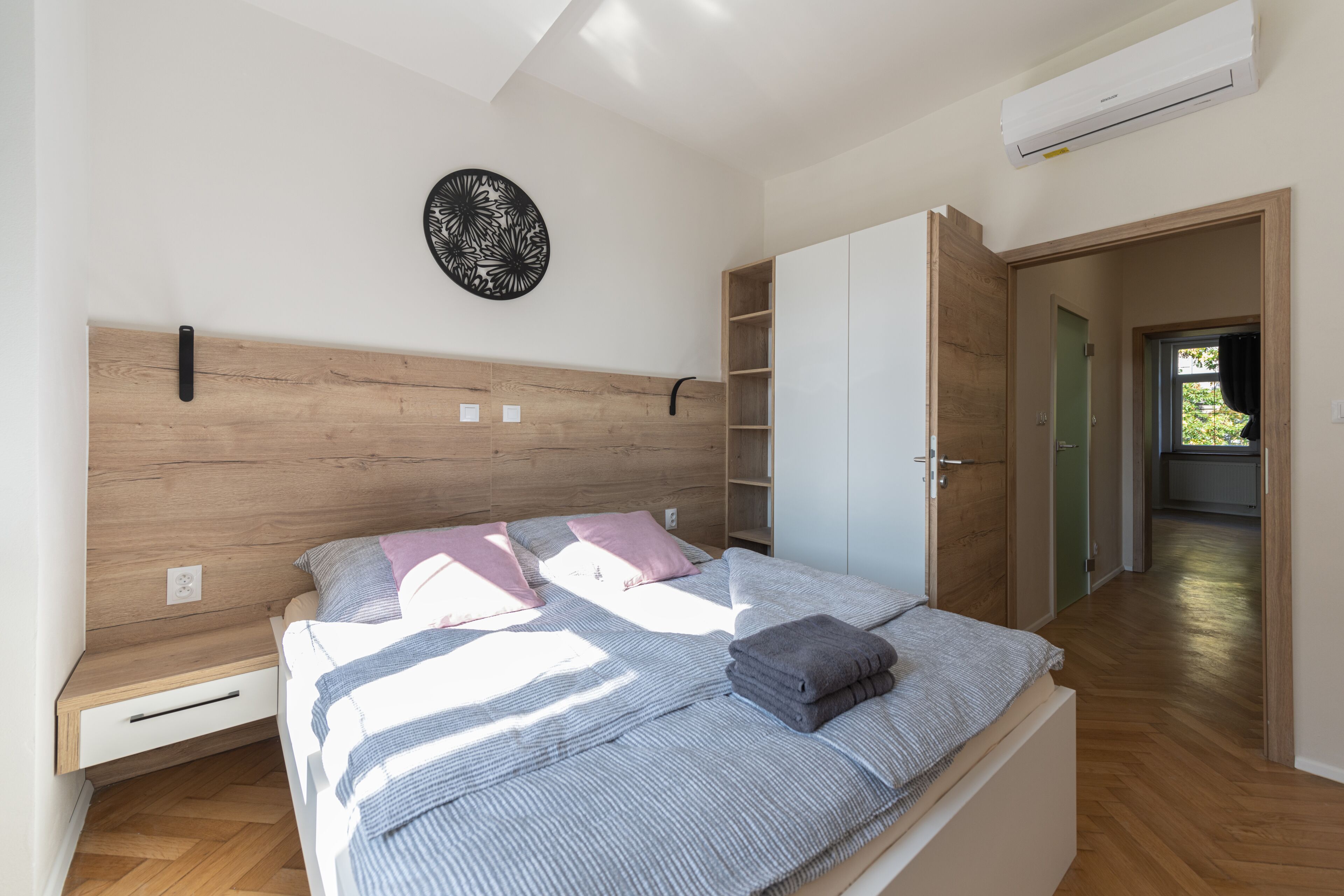 A bedroom with a wooden headboard, a bed with striped bedding, a bedside table, a wall-mounted air conditioner, and a door leading to another room.