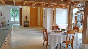 Un comedor con una mesa dispuesta para cuatro personas, sillas de madera, mantel blanco y vistas a una cocina y una puerta que da al exterior.