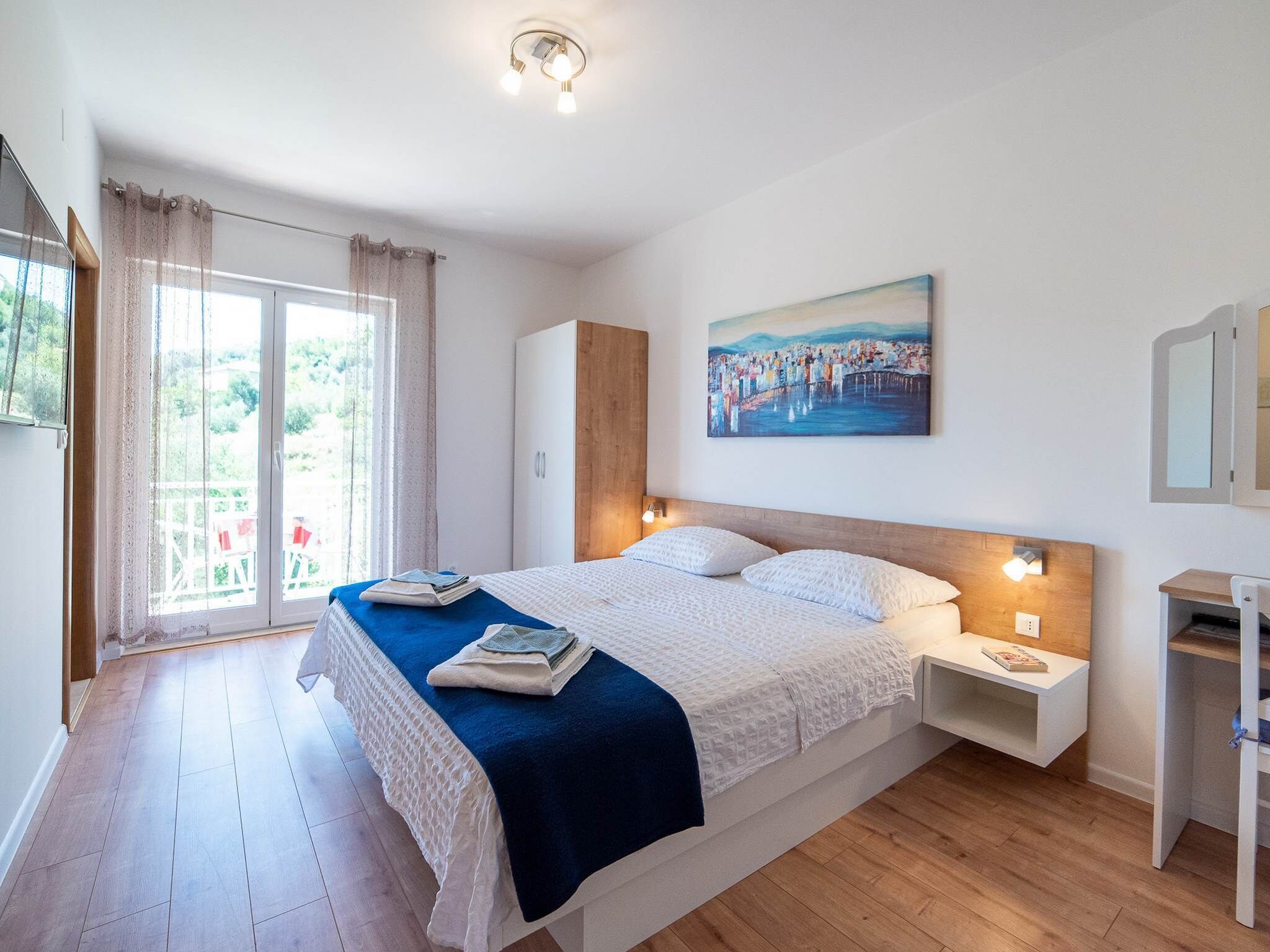 Une chambre à coucher avec un grand lit, une table de chevet, un bureau, un tableau et une vue sur l’extérieur.