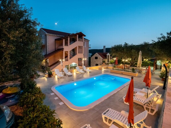 A swimming pool with lounge chairs and umbrellas, surrounded by a building with a balcony and a stone structure. - Apartments Njirić  (Dubrovnik)