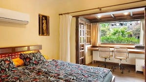 Une chambre avec un lit, un bureau avec deux chaises, une fenêtre donnant sur un espace verdoyant et un climatiseur fixé au mur.