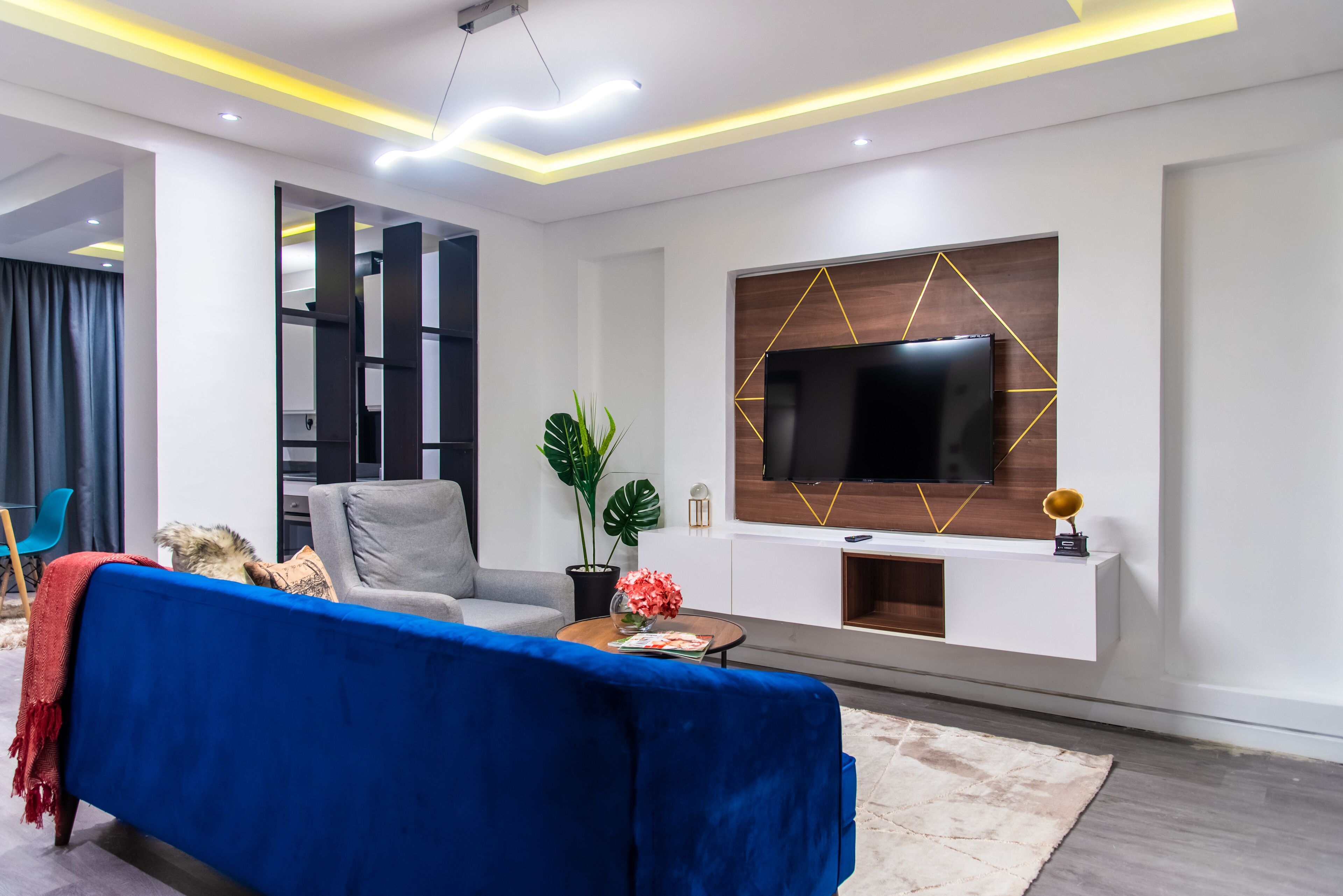 A modern living room with a blue sofa, grey armchair, and a large flat-screen TV mounted on a wooden wall.