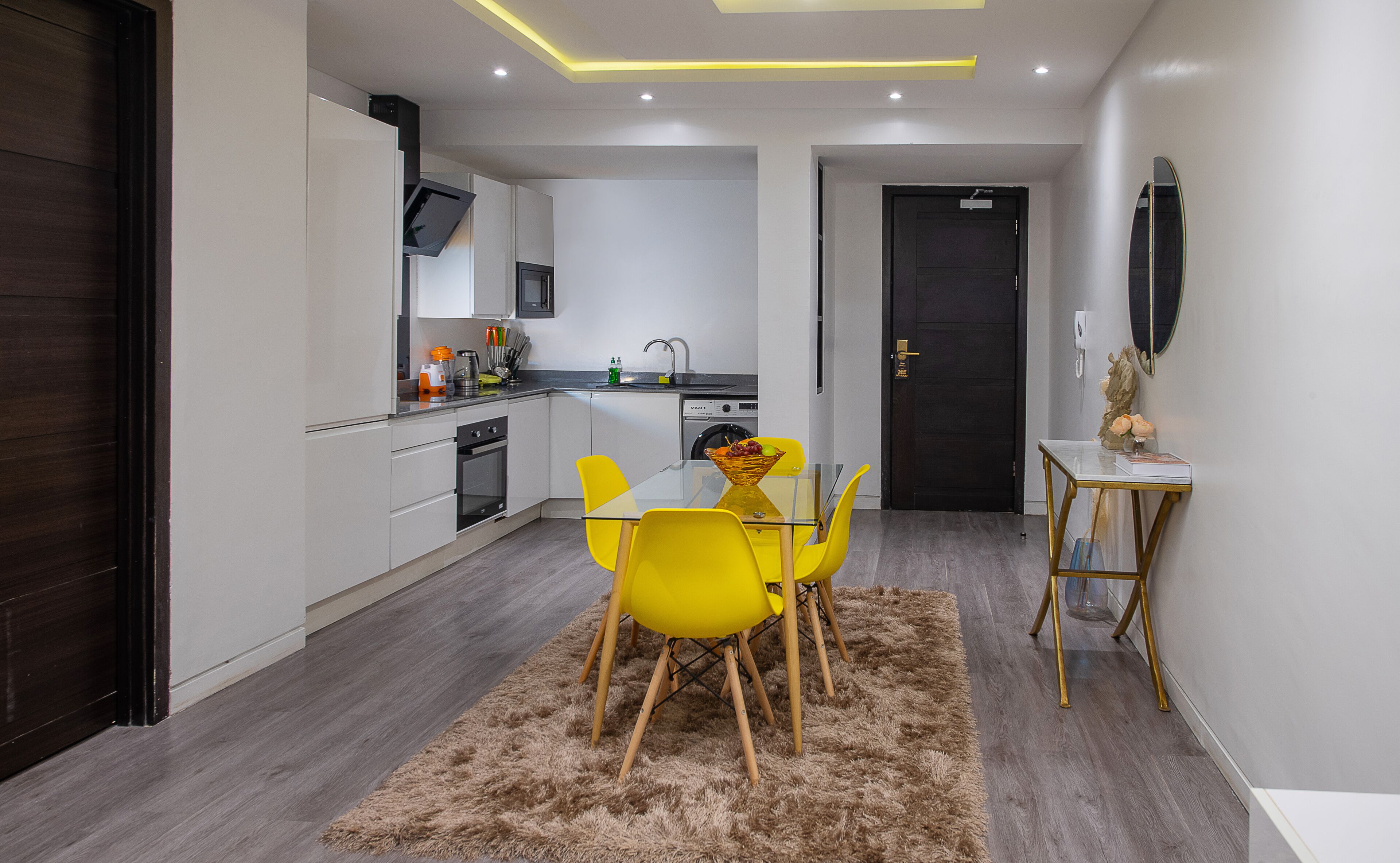 A modern kitchen with white cabinets, a built-in oven, and a dining area with a glass table and yellow chairs.