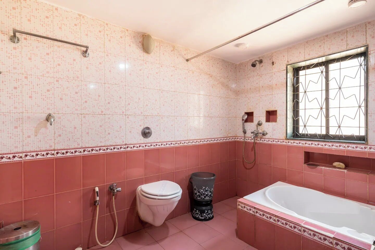 Une salle de bain avec des murs recouverts de carreaux roses, des toilettes blanches, une poubelle noire, une baignoire et une fenêtre avec une grille métallique.