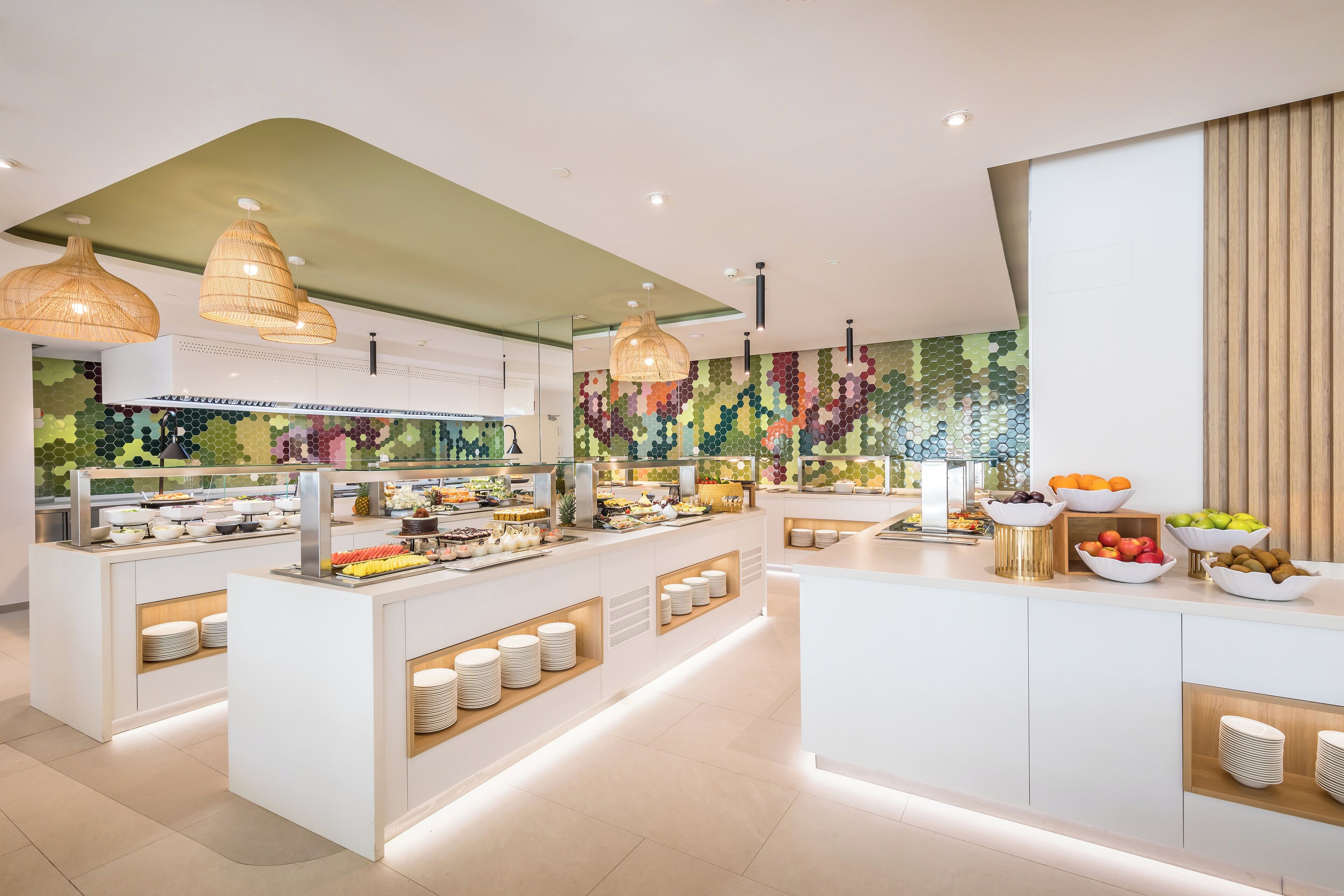 A modern breakfast bar with a variety of food options, including fruits and pastries, and a colorful mosaic wall in the background.