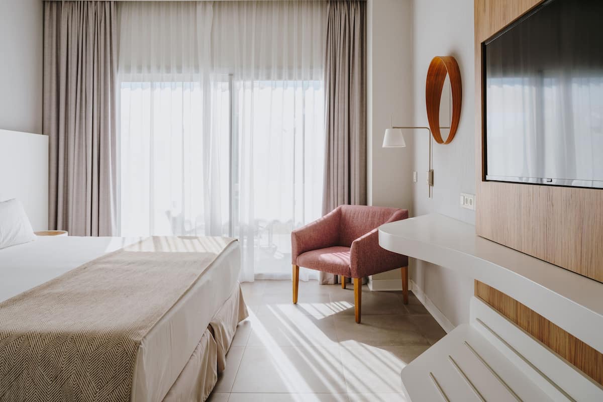 Habitació d'hotel amb llit, una cadira, televisió i una finestra gran amb cortines.