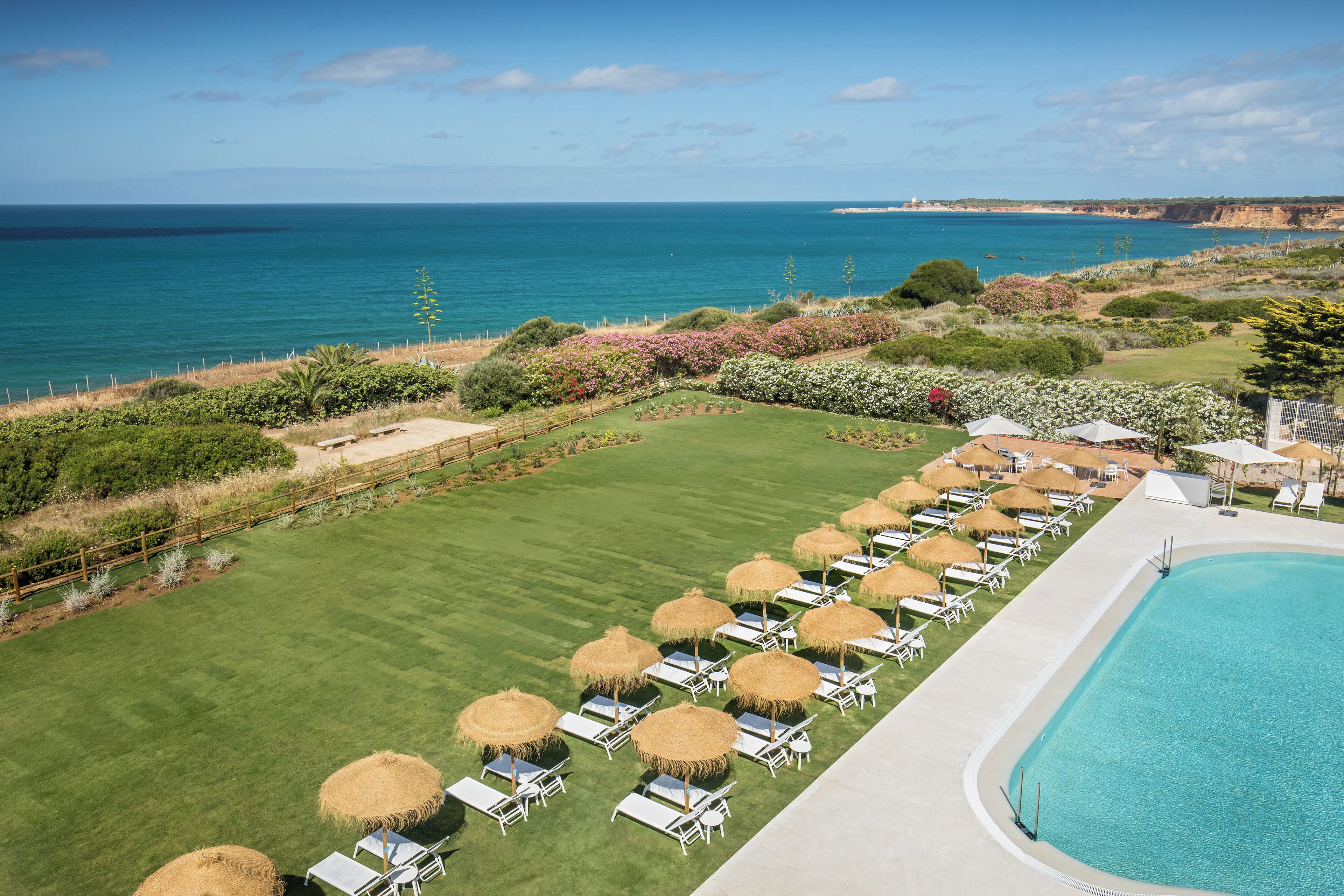 Zona de piscina con sillones reclinables y sombrillas, césped cuidado y vista al océano.
