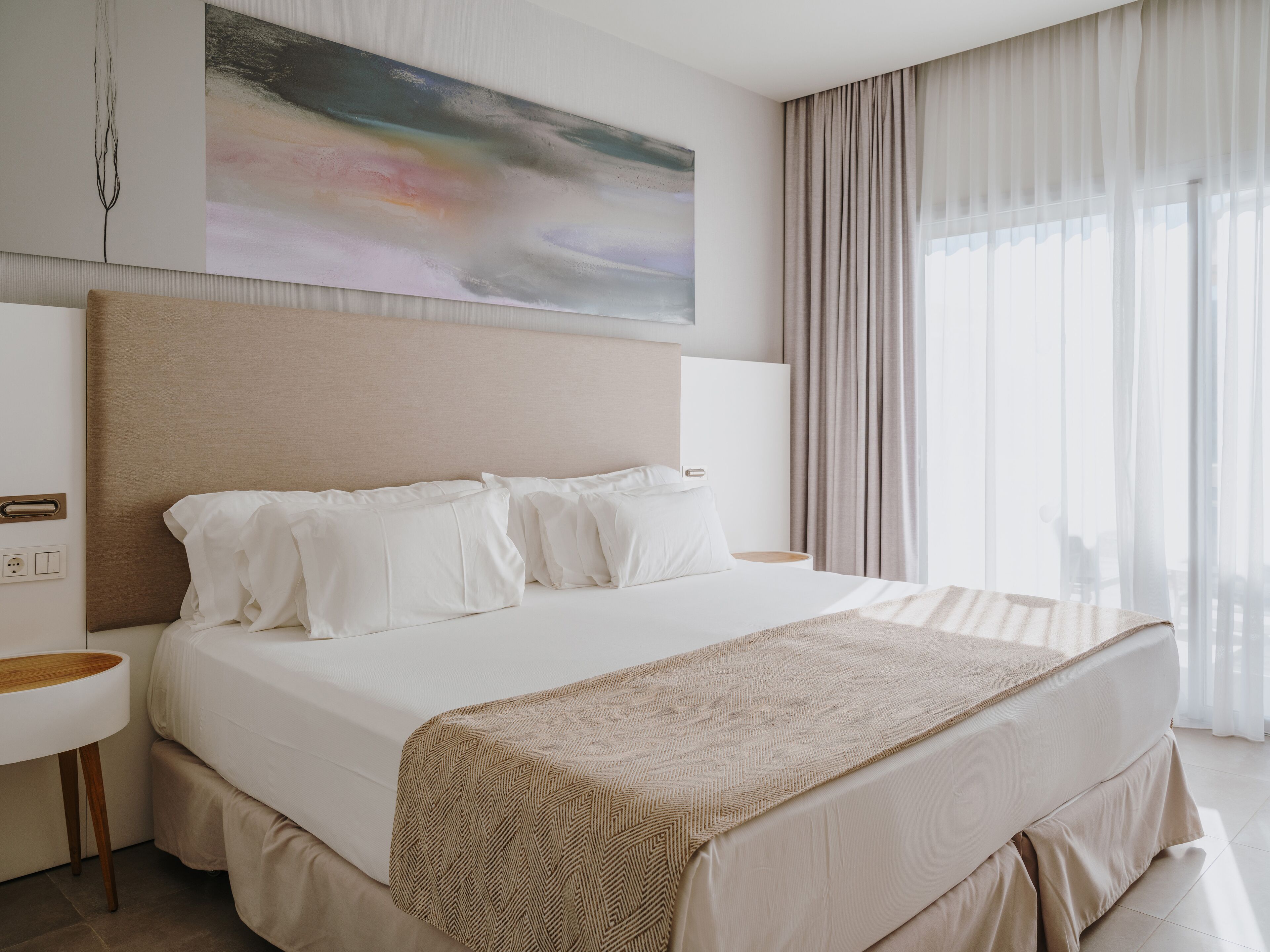 Una habitación de hotel moderna con una cama grande, una mesita de noche, una obra de arte en la pared y un amplio ventanal con cortinas.