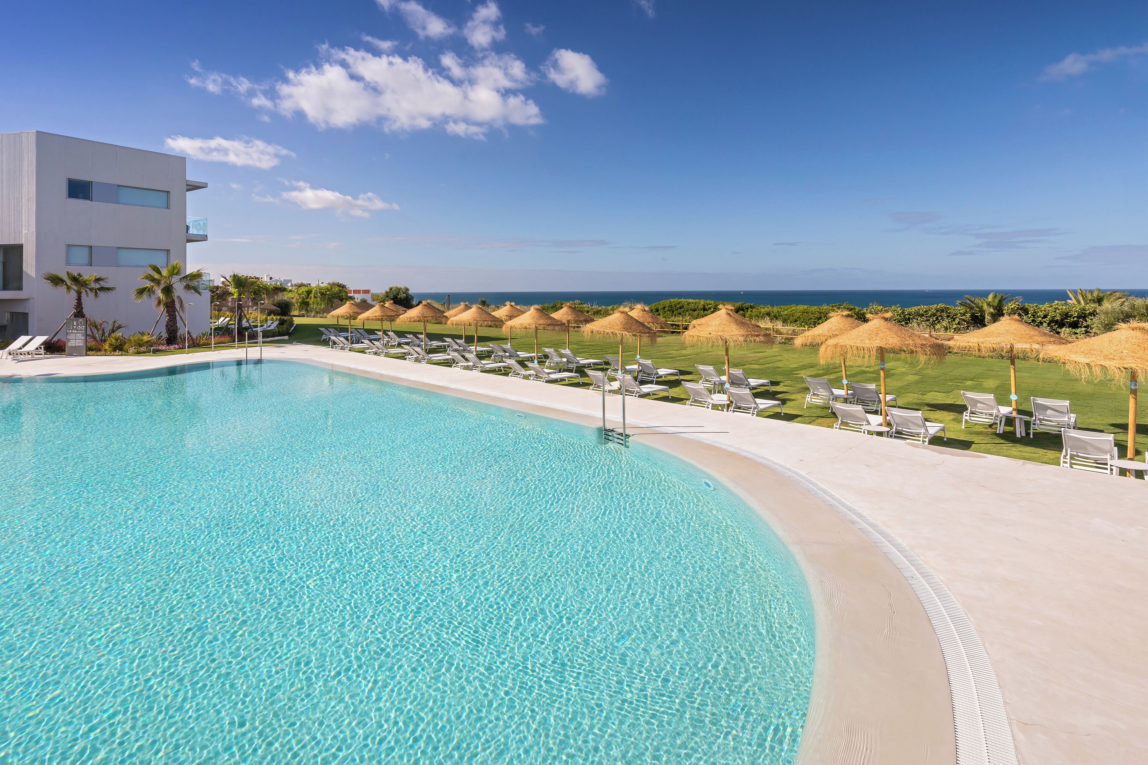 Une piscine à l’eau d’un bleu cristallin, entourée de chaises longues et de parasols, avec vue sur l’océan et un bâtiment moderne en arrière-plan.