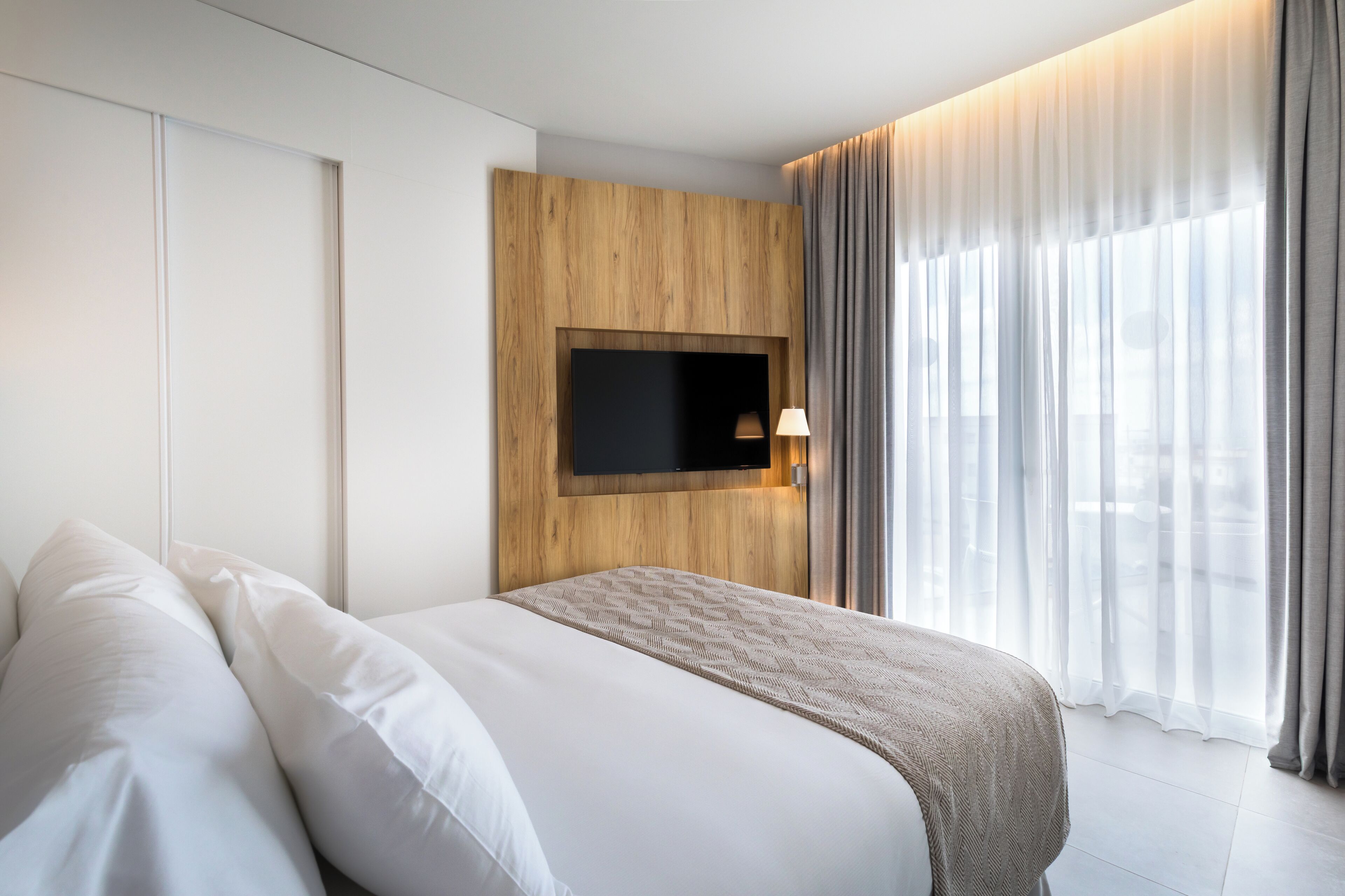 Ein modernes Hotelzimmer mit einem großen Bett, einem wandmontierten Fernseher und Blick auf die Stadt durch transparente Vorhänge.