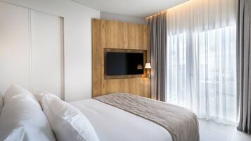 A modern hotel room with a large bed, a mounted TV, and a view of the city through sheer curtains.