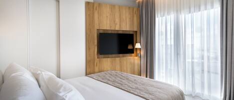 Habitación de hotel moderna con una cama grande, un televisor montado y vistas a la ciudad a través de cortinas de gasa.