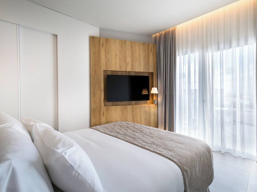 A modern hotel room with a large bed, a mounted TV, and a view of the city through sheer curtains.