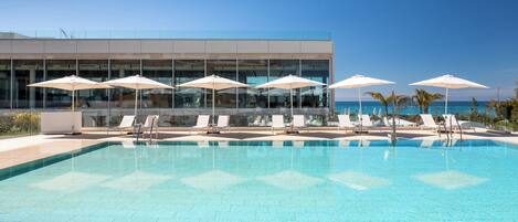 A clear blue pool with lounge chairs and umbrellas, a modern building with large windows, and a clear sky.