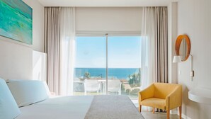 A hotel room with a bed, a yellow armchair, a large window with a view of the sea, and a painting on the wall.