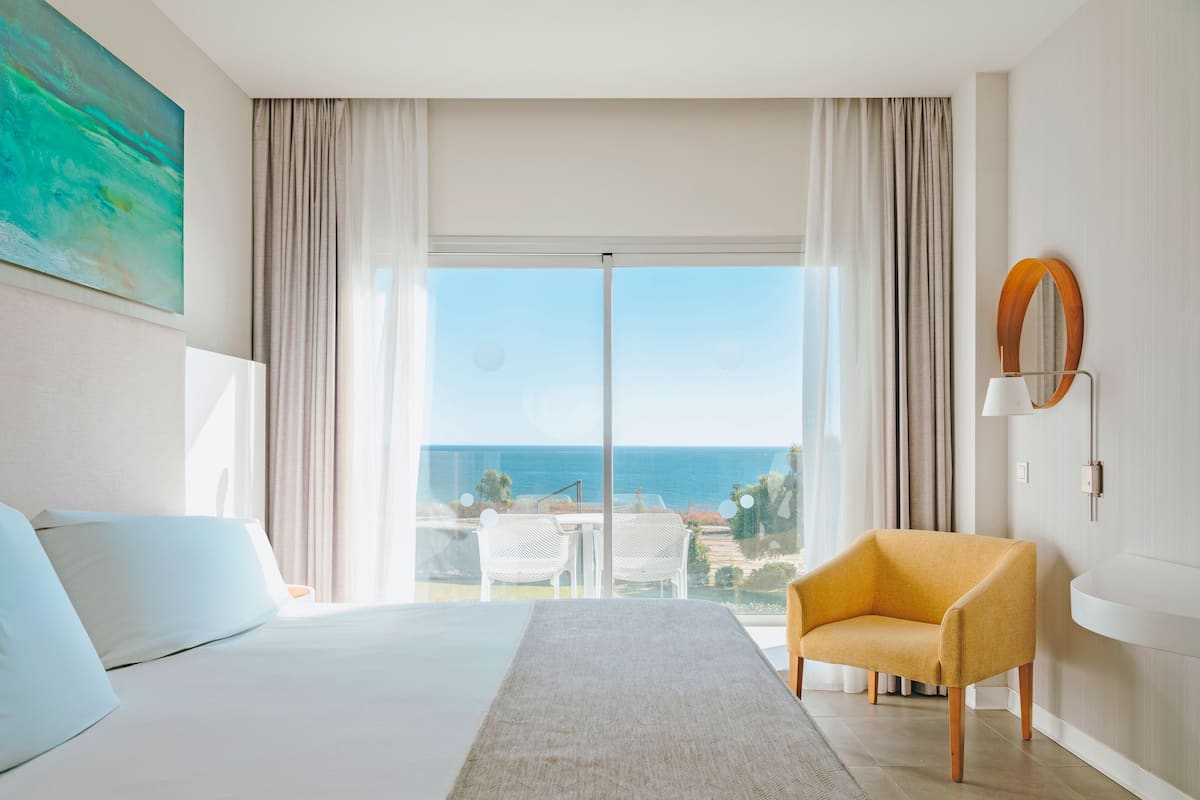Habitació d'hotel amb un llit, un butacó groc, una finestra gran amb vista al mar i un quadre a la paret.