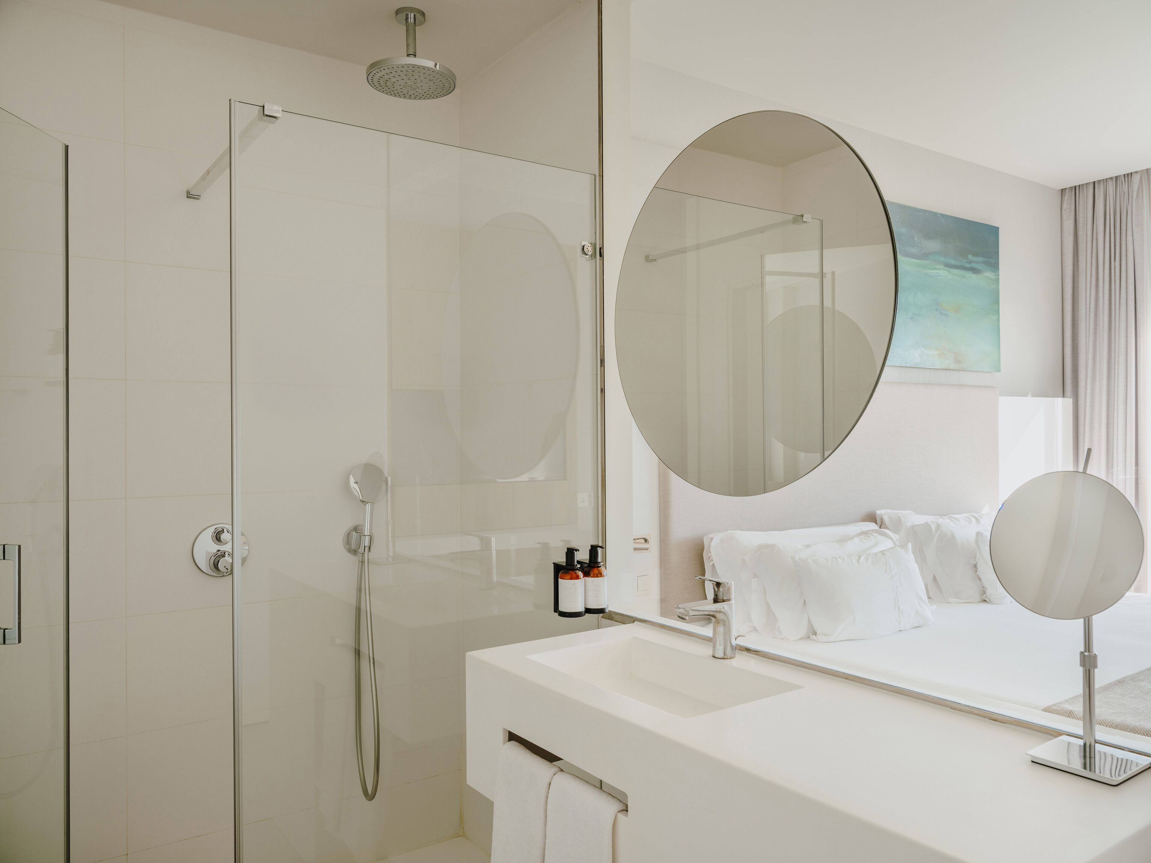 Un baño moderno con ducha envuelta en vidrio, un espejo redondo grande y un lavabo blanco.
