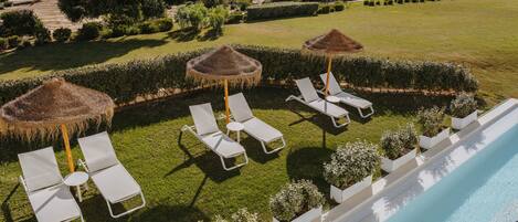 A poolside area with lounge chairs and a thatched umbrella, surrounded by well-manicured lawns and a view of the sea.