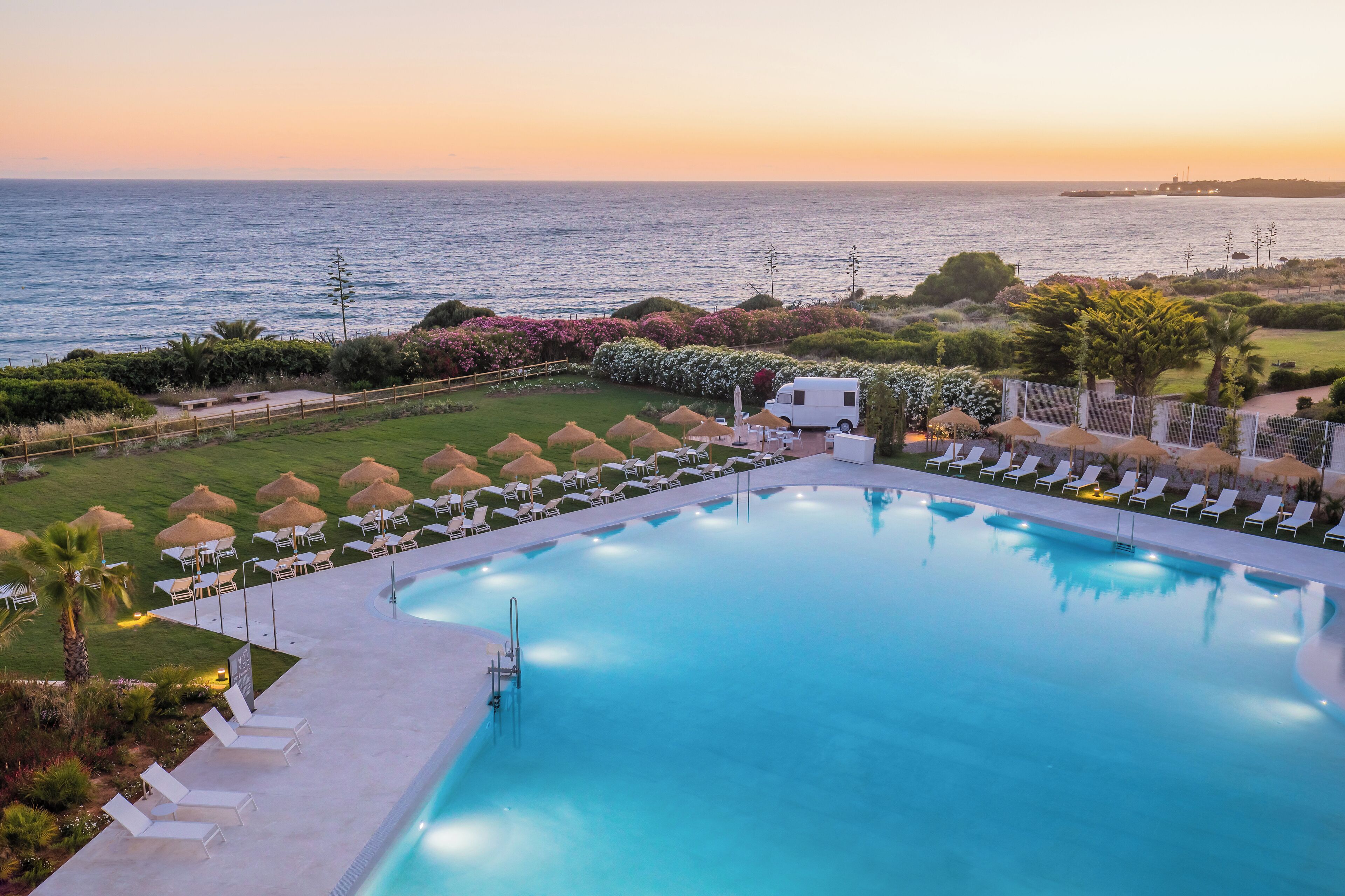A pool with lounge chairs and umbrellas, surrounded by greenery and a view of the sea at sunset.