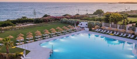A pool with lounge chairs and umbrellas, surrounded by greenery and a view of the sea at sunset.