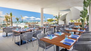 An outdoor dining area with tables and chairs, umbrellas, and a view of the ocean.