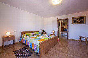 Un dormitorio con una cama de madera, una mesita de noche con lámpara, un taburete pequeño y una puerta que da a otra habitación.