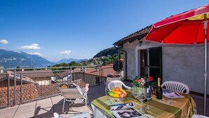 A terrace with a table set for dining, a red umbrella, and a view of distant mountains. - Terrazza Vista Menaggio (Plesio)