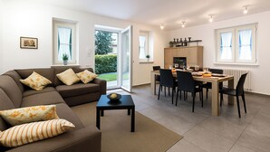 A modern living room with a sofa, a dining table, and chairs. There is a kitchen area with cabinets and a window. - Lenno Dolcelago (Tremezzina)