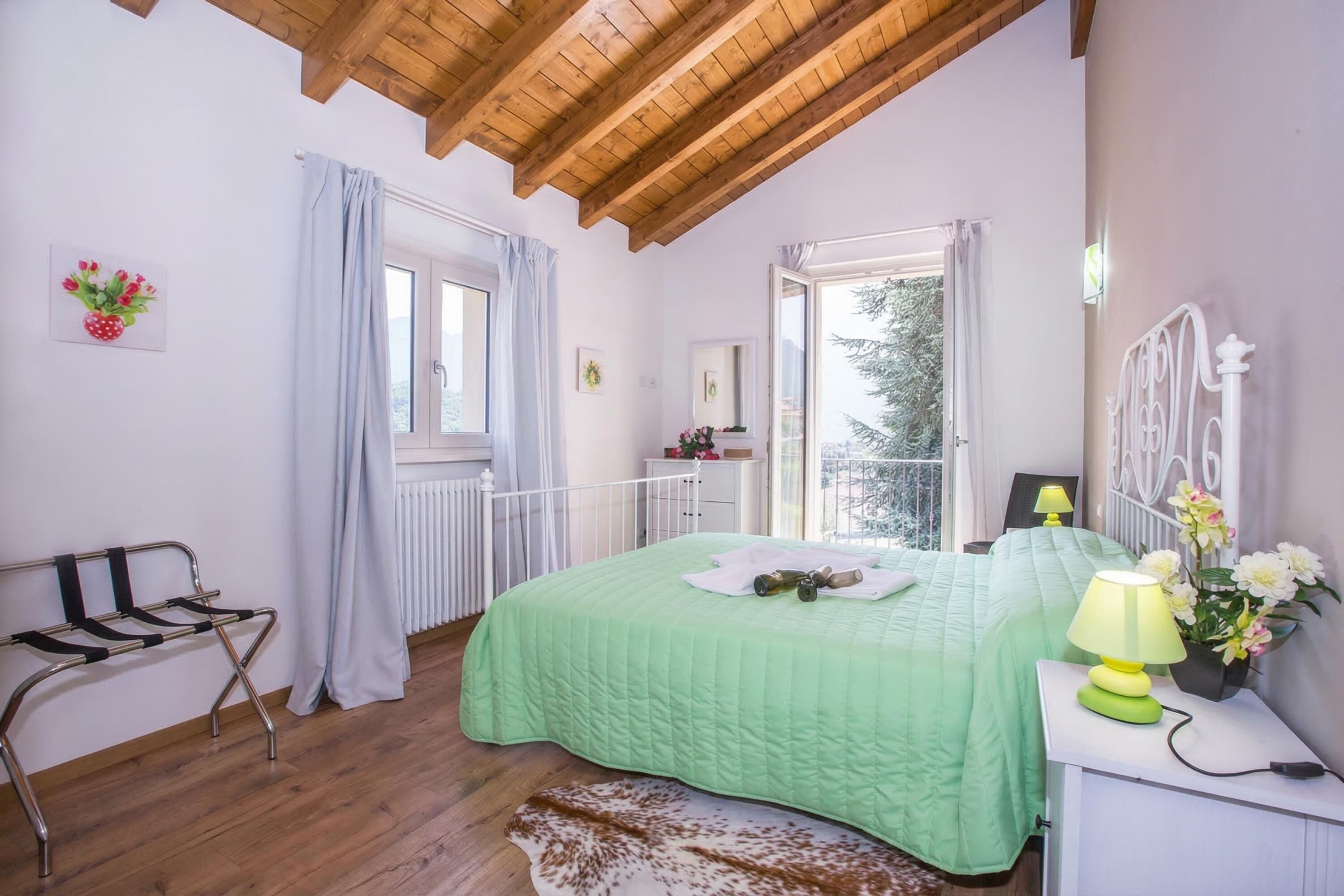 A bedroom with a green bedspread, a bedside lamp, a radiator, a window with curtains, and a view of trees outside.