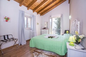 A bedroom with a green bedspread, a wooden ceiling, a bench, a bedside table with a lamp, and a window with curtains.