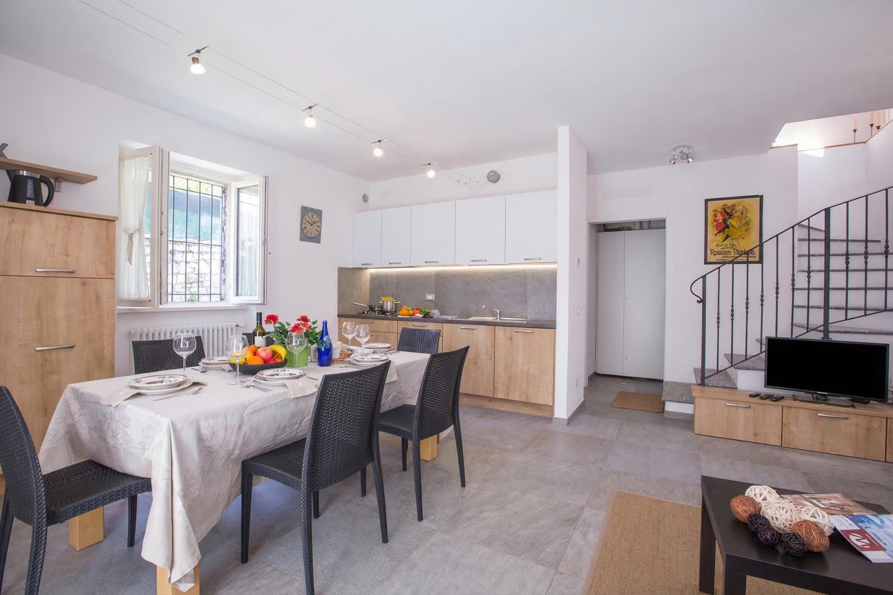 Una cocina moderna con armarios blancos, un comedor con una mesa dispuesta para cuatro personas y una escalera con un televisor.