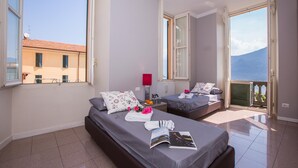 A hotel room with two beds, a balcony with a view of the sea, and a window overlooking a building. - Casa Daniele (Menaggio)