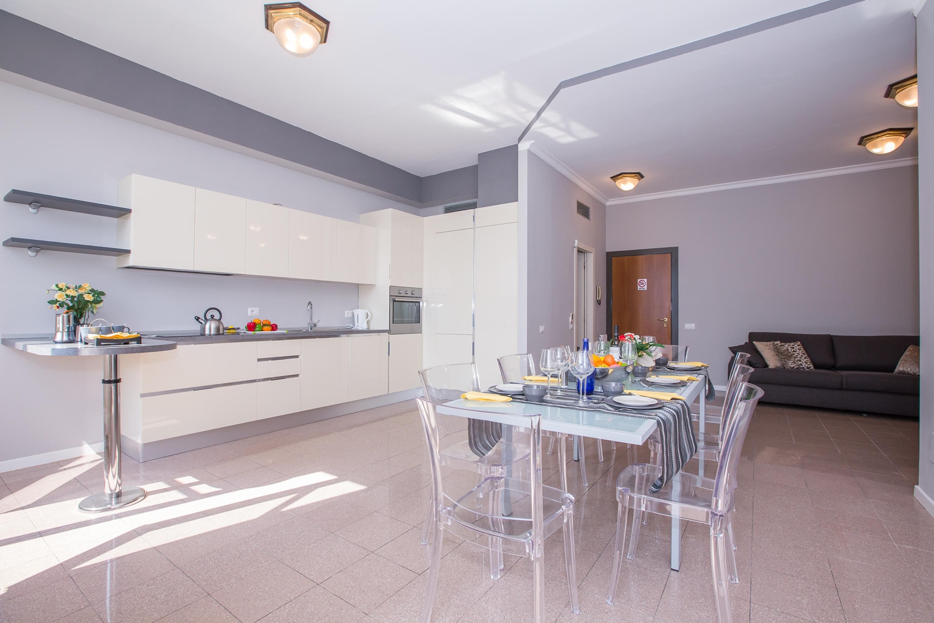 A modern kitchen with white cabinets, a dining area with clear chairs, and a grey sofa.
