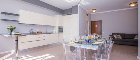 A modern kitchen with white cabinets, a dining area with clear chairs, and a grey sofa.