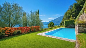 A rectangular swimming pool with a stone border, surrounded by well-maintained grass and a hedge with red flowers. - Villa San Michele (Griante)
