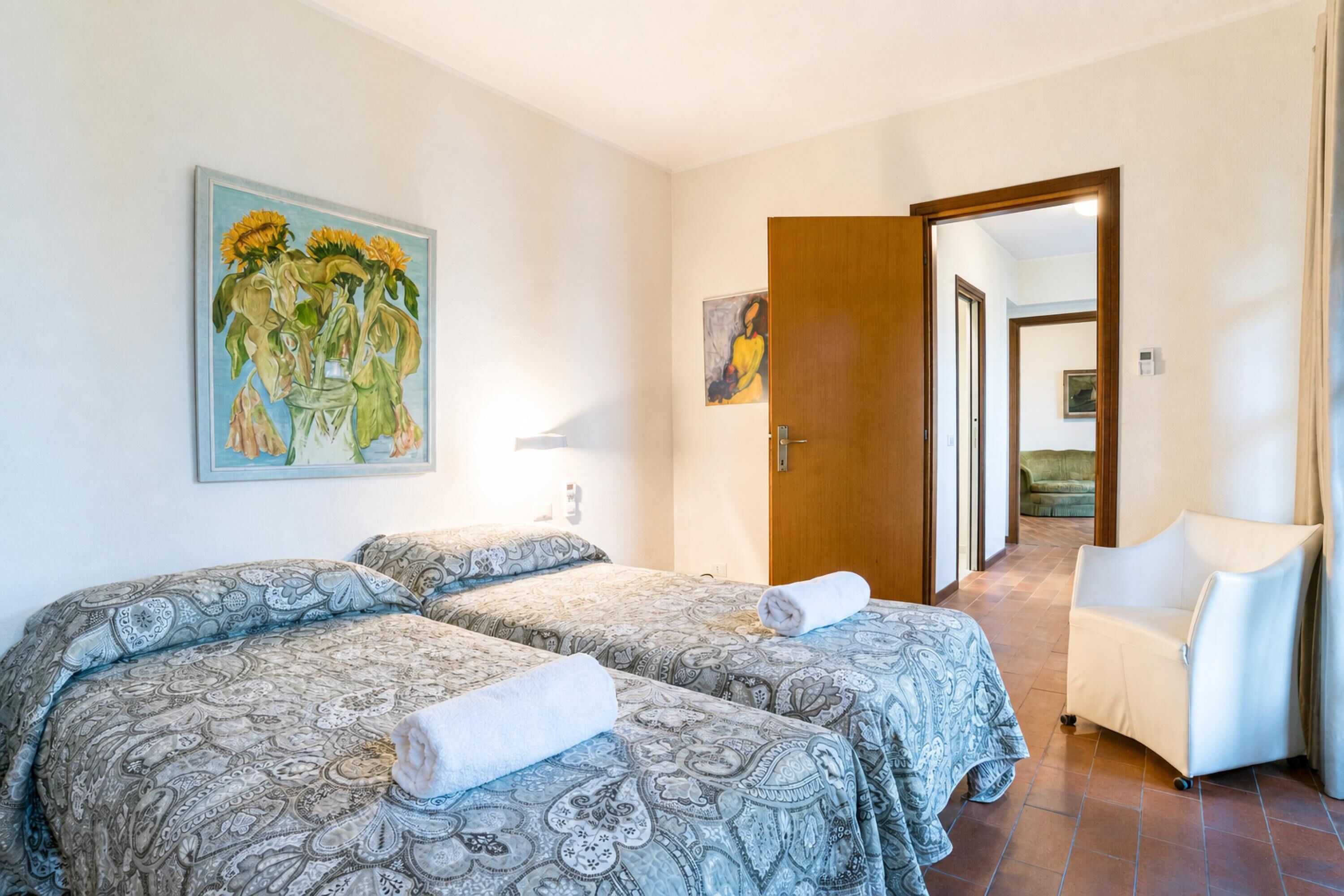Een hotelkamer met twee bedden, een zonnebloemschilderij, een stoel en een deur naar een andere kamer.