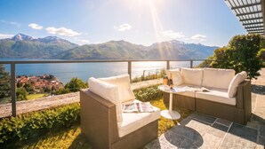 A terrace with wicker furniture, a table with a bottle and glasses, and a view of a lake and mountains. - Penthouse-LakeView-AC-PrivateGarden (Menaggio)