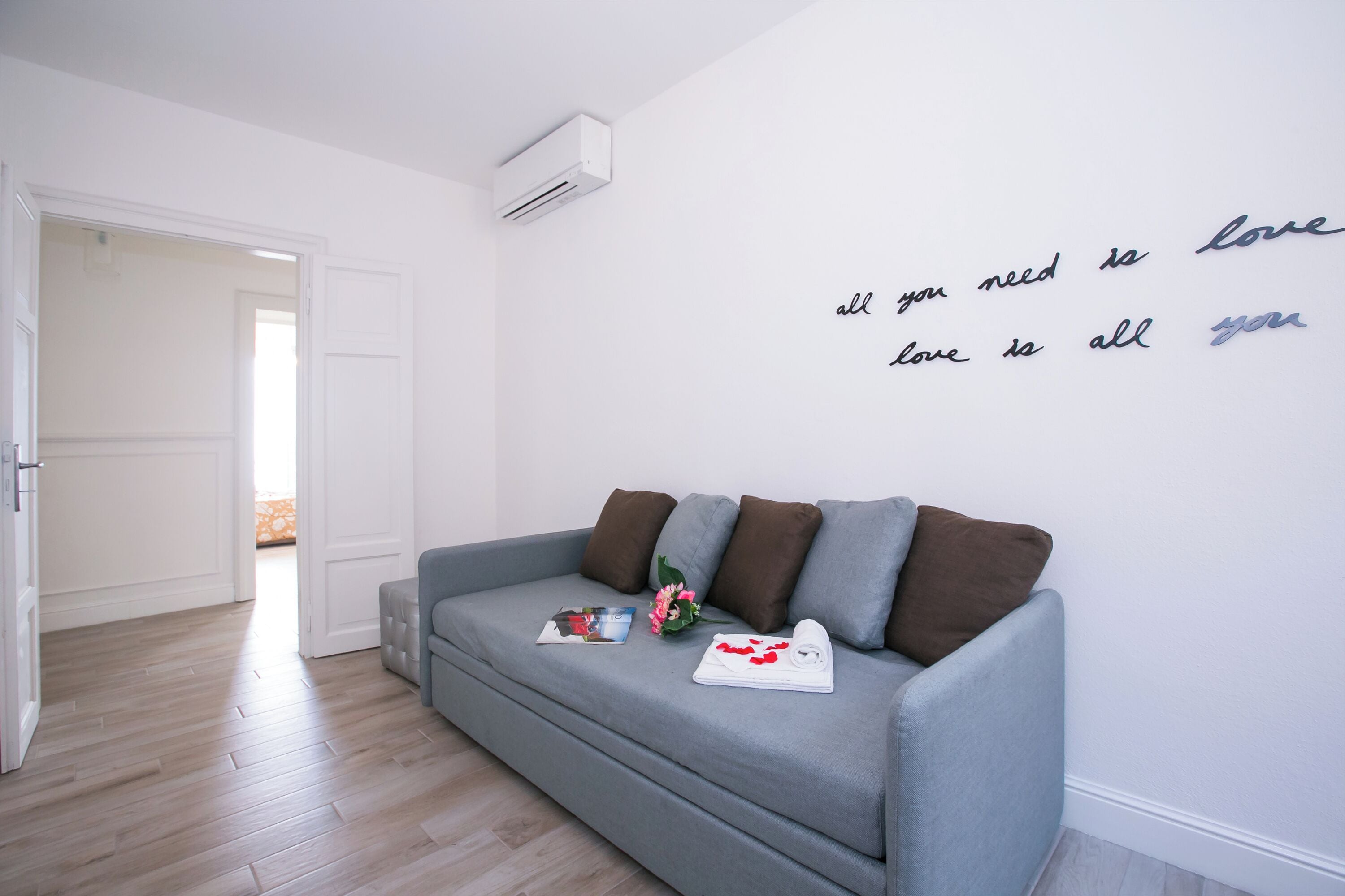 A modern living room with a grey sofa, a wall with a love quote, and a door leading to another room.