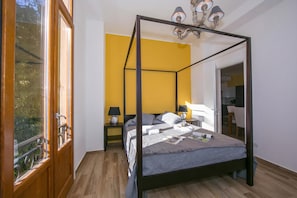 A four-poster bed with a wooden frame, a nightstand with a lamp, and a view through a window with a wooden railing. - Villa Evelyn Terrazzo (Menaggio)