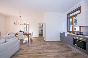 A modern kitchen with a built-in oven, a dining area with a wooden table and chairs, and a living area with a sofa and a chandelier. - Villa Evelyn Terrazzo (Menaggio)