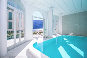 A modern indoor swimming pool with clear glass doors leading to a terrace with a mountain view. - Villa Larius Balcone (Carate Urio)