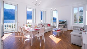 A spacious living area with a dining table, white furniture, a sofa, and a television. - Villa Larius Balcone (Carate Urio)