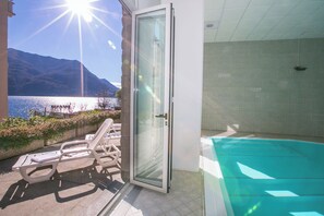Un balcone con una porta vetrata che si apre su una piscina, una sedia a sdraio e vista su un lago e montagne.