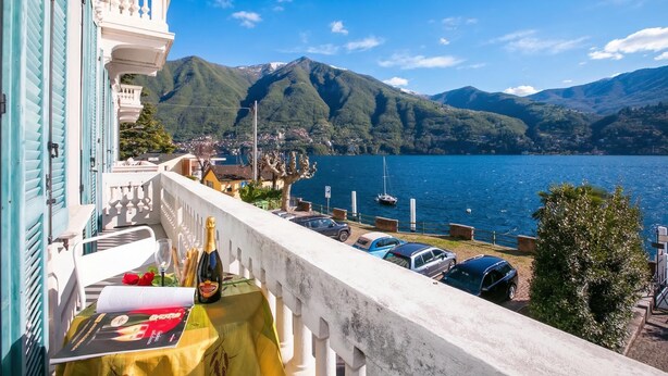 A balcony with a table set for a meal, overlooking a lake with boats and mountains in the background. - Villa Larius Balcone (Carate Urio)