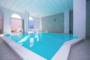 Una amplia piscina interior con agua cristalina y azul, rodeada de paredes blancas y suelo de baldosas.