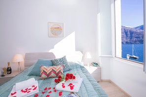 A bedroom with a bed, pillows, a nightstand, a lamp, a framed picture, and a view of a body of water through a window. - Villa Larius Balcone (Carate Urio)