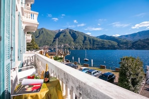 A balcony with a table set for a meal, overlooking a lake with boats and mountains in the background. - Villa Larius Balcone (Carate Urio)